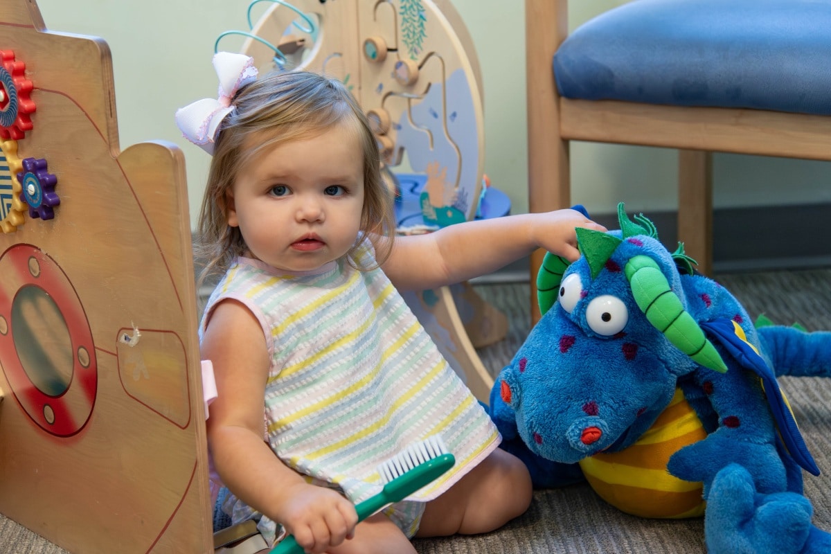 Toddler playing with a toothbrush and stuffed dragon toy at Wilmington Pediatric Dentistry.