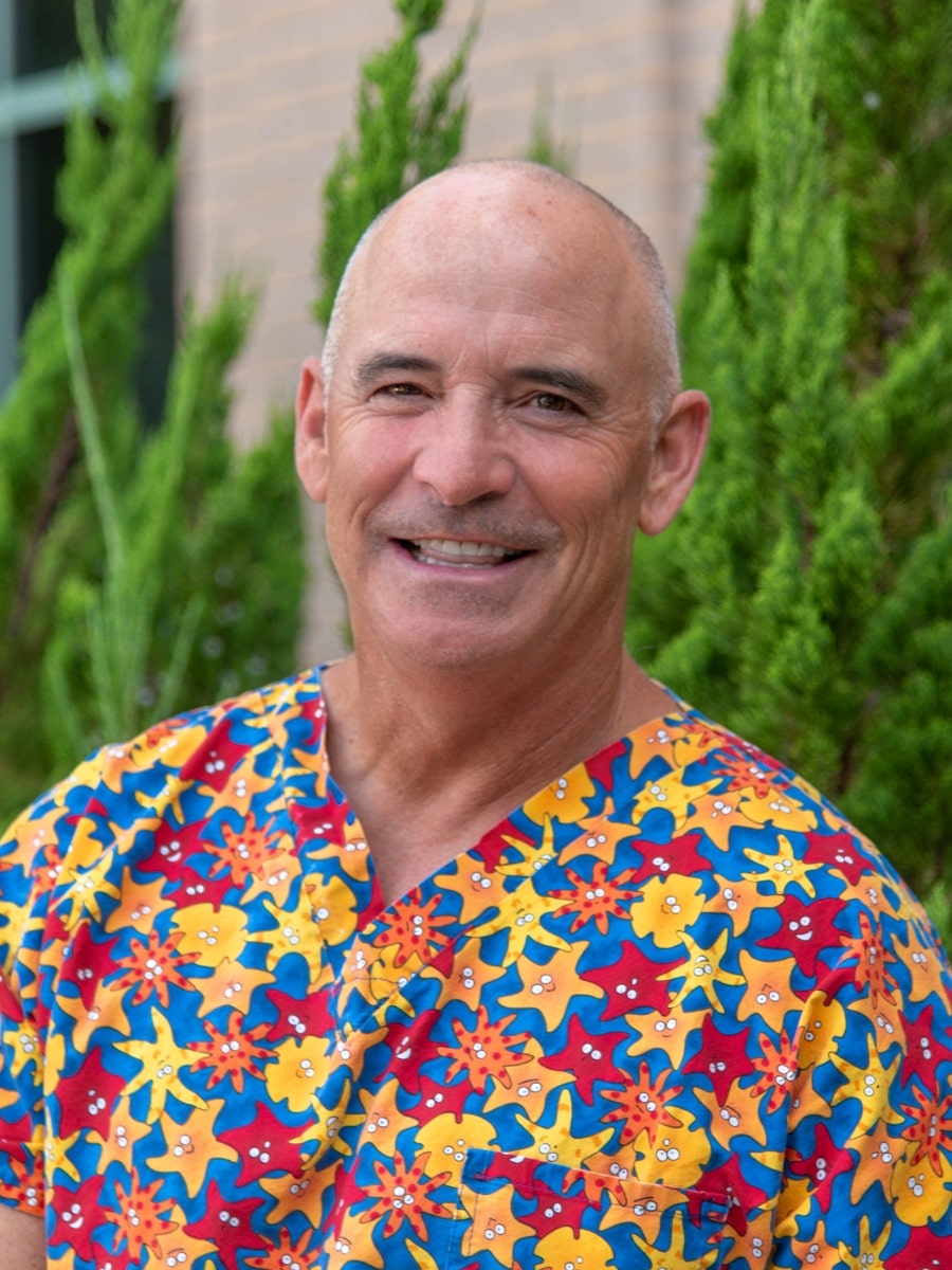Smiling male dental professional in colorful scrubs standing outdoors in front of greenery